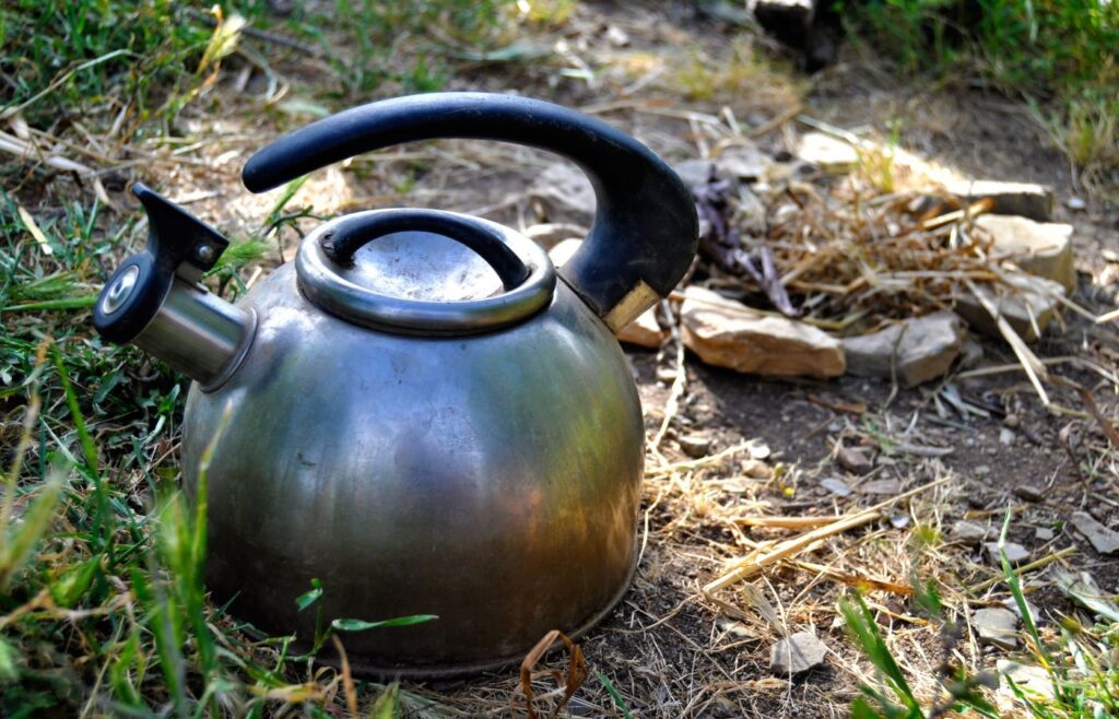 An image of a kettle outdoors in the mud, surrounded by sticks and grass. As if being used for outdoor activities with children.