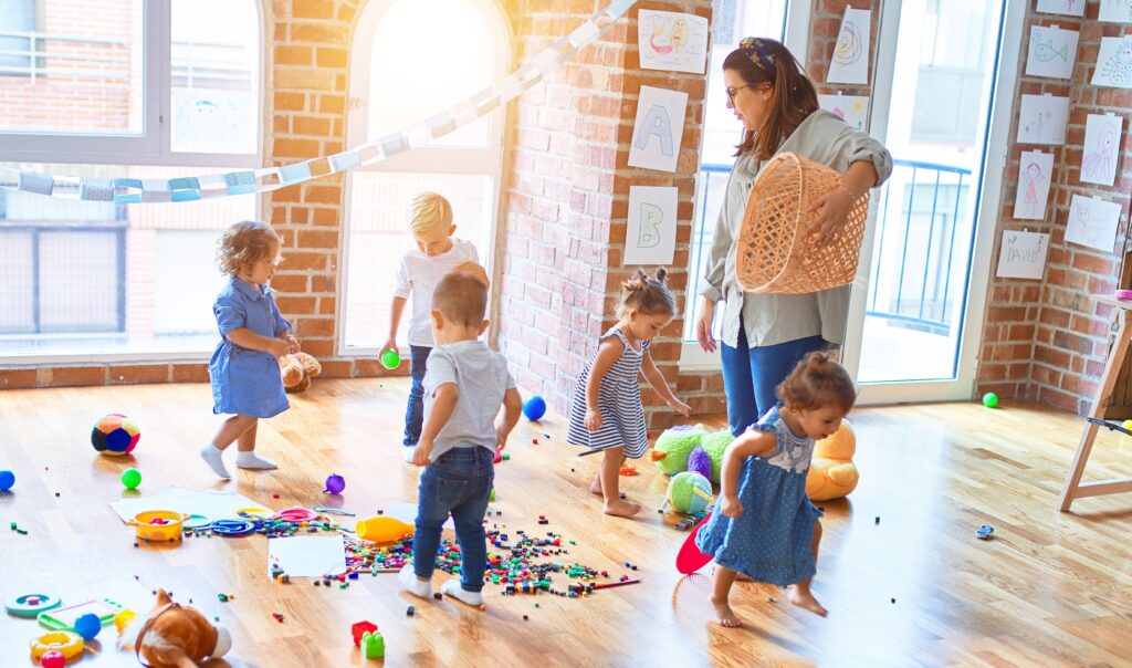 Female nursery practitioner stood holding a basket surrounded by toddlers playing with abstract toys.