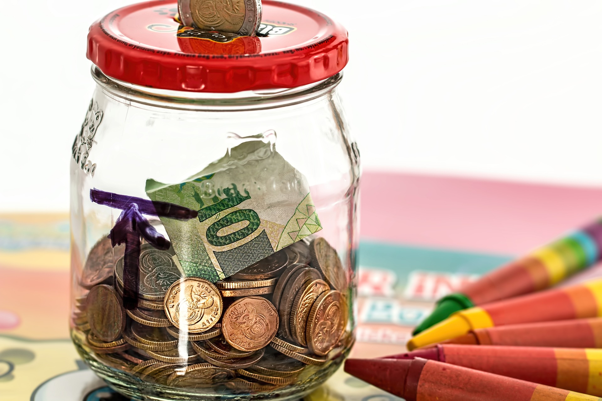 A glass jar with a red lid containing money, as if a piggy bank. There are multi-colour crayons to the right of the jar.