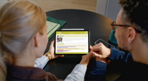 The back of a woman and a man who are sat in front of a low black table. They're looking at a tablet with the eylog learning journal on the screen.